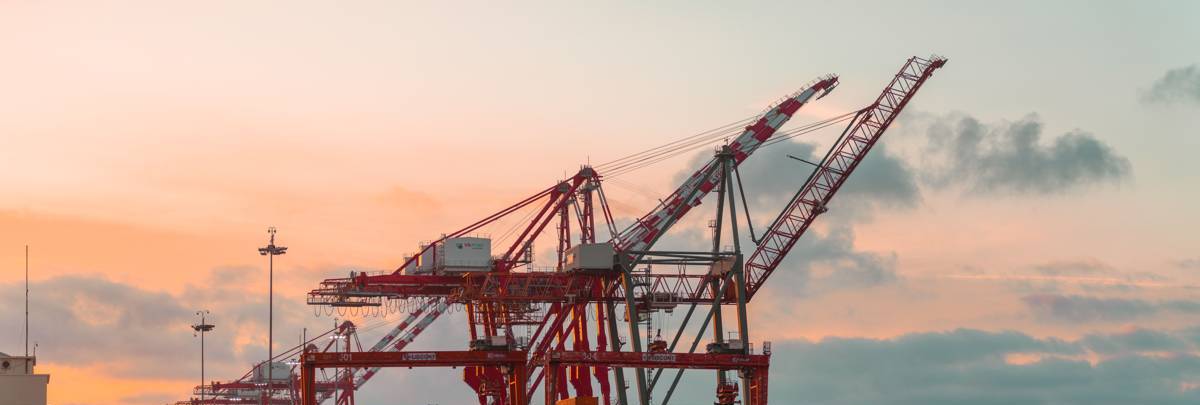 Red container cranes silhouetted against a pastel sky at a port, generic illustrative photograph