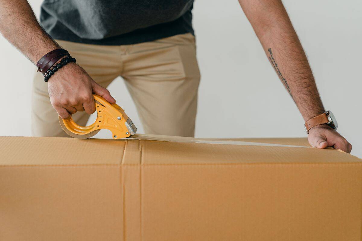 Person using a yellow tape dispenser to seal a plain cardboard carton.